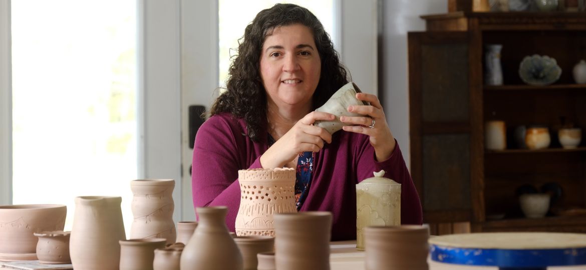 Photo of 2023 ASC Emerging Creators Fellowship recipient Maria Frey. She's holding a piece she fired using a soda kiln, at a workshop. In the foreground are works in progress, yet to be fired.
