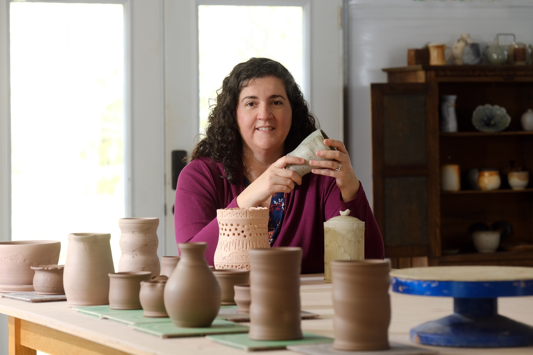 Photo of 2023 ASC Emerging Creators Fellowship recipient Maria Frey. She's holding a piece she fired using a soda kiln, at a workshop.  In the foreground are works in progress, yet to be fired.
