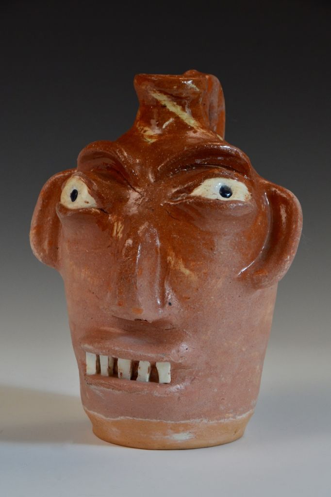 This face jug by Burlon Craig features bulging eyes, five buck teeth, red clay/reddish/pink glaze and black pupils.